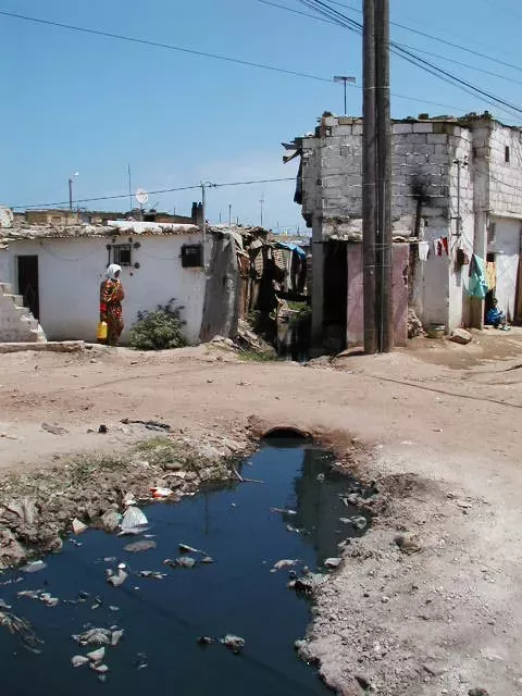 Dilapidated buildings with exposed wiring and laundry hanging outside; a person in colorful clothing walks by, and a pool of stagnant water with trash is in the foreground, capturing a slice of life in Tunisia.