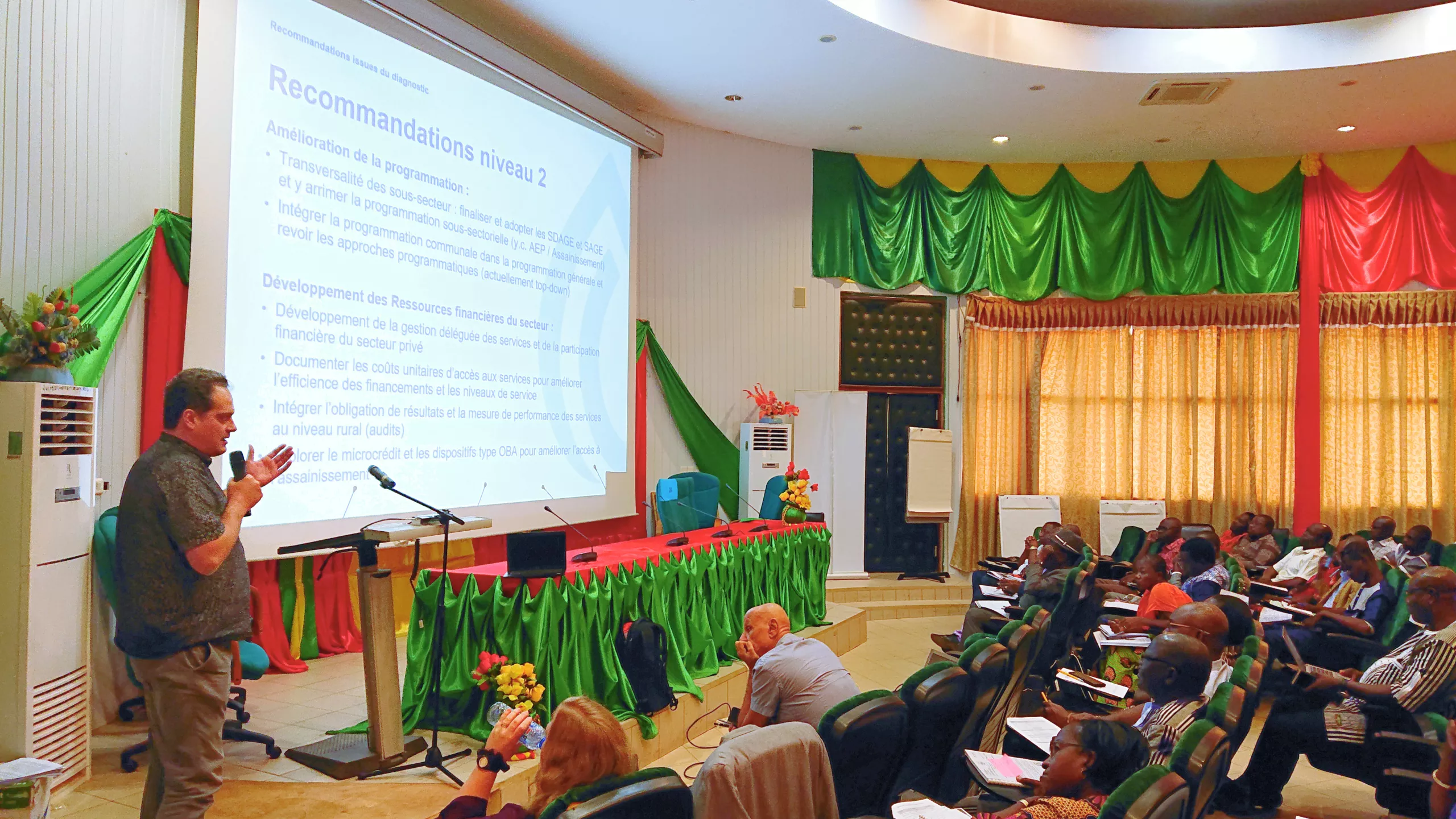 A man presents at a conference with a slide titled "recommendations in area 2" behind him; attendees, seated at tables, listen attentively in a decorated hall.