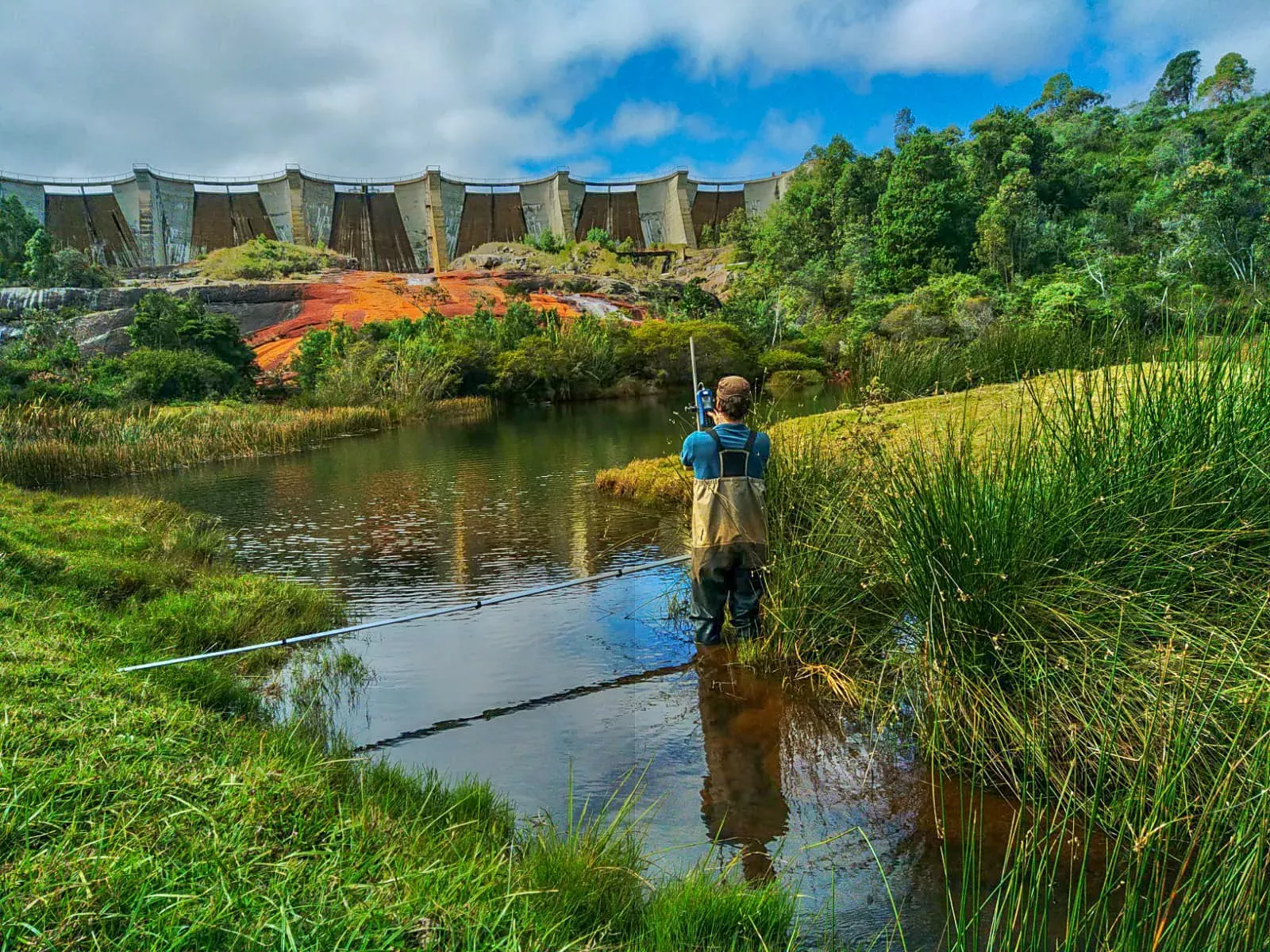 Man in waders fishing in a river with a large dam in the background, surrounded by lush greenery under a cloudy sky.