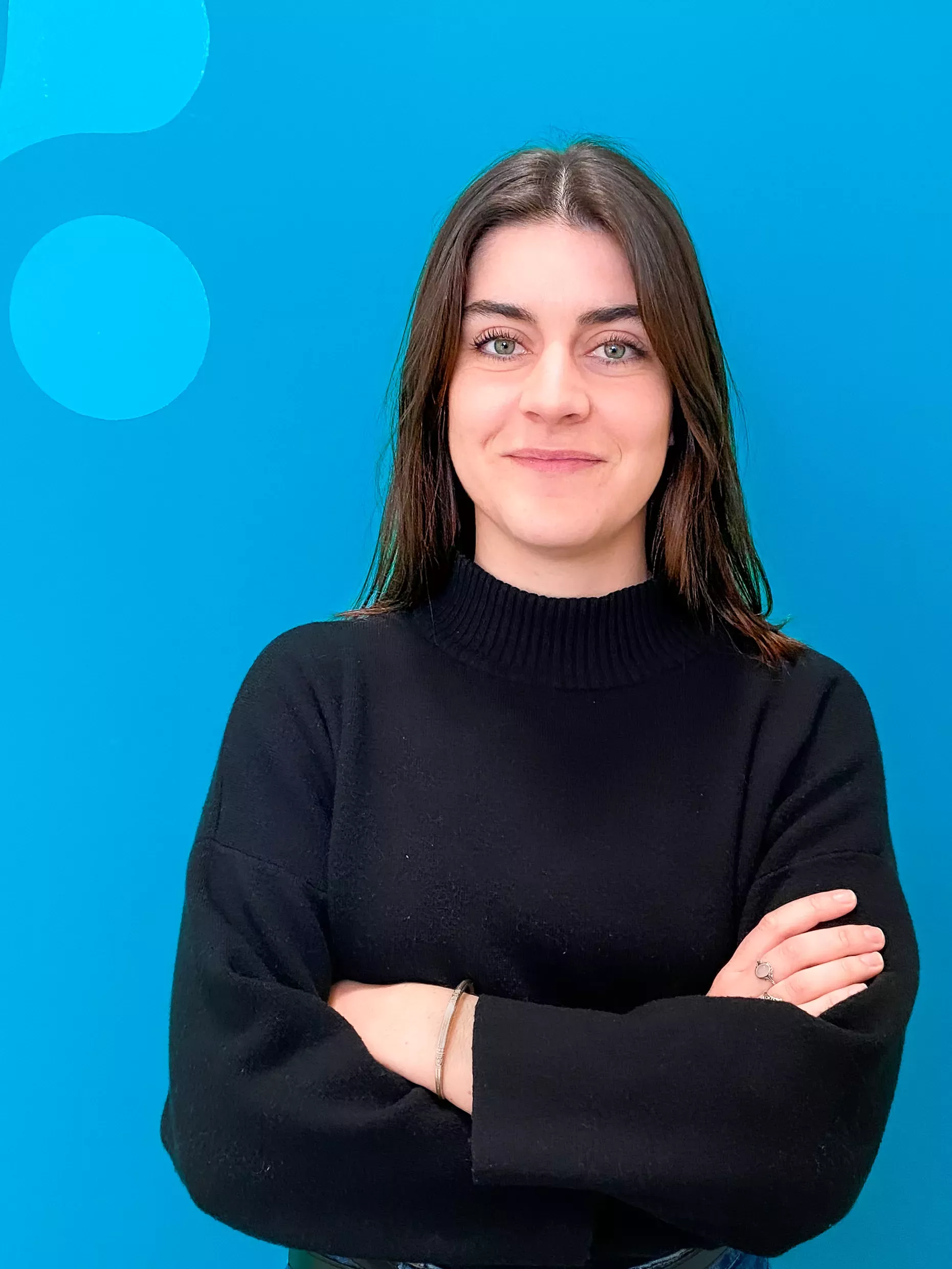 Woman with dark hair, wearing a black sweater, standing against a blue background, arms crossed, smiling.