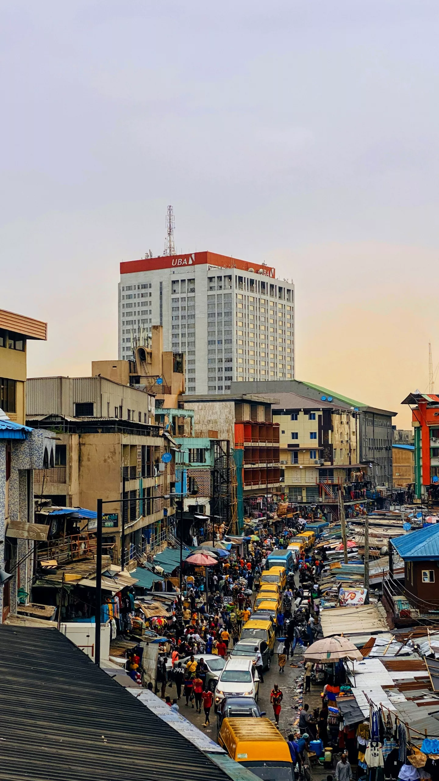 View of a bustling market with colorful stalls under umbrellas, foregrounding a taller urban backdrop with a prominent uba building.