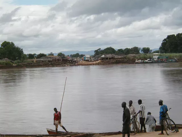 Several people, some with bicycles, gather near a calm river with a small boat in the foreground. In the background, there are trees, a village reminiscent of those in Irak, and an institutional truck parked near the riverbank.