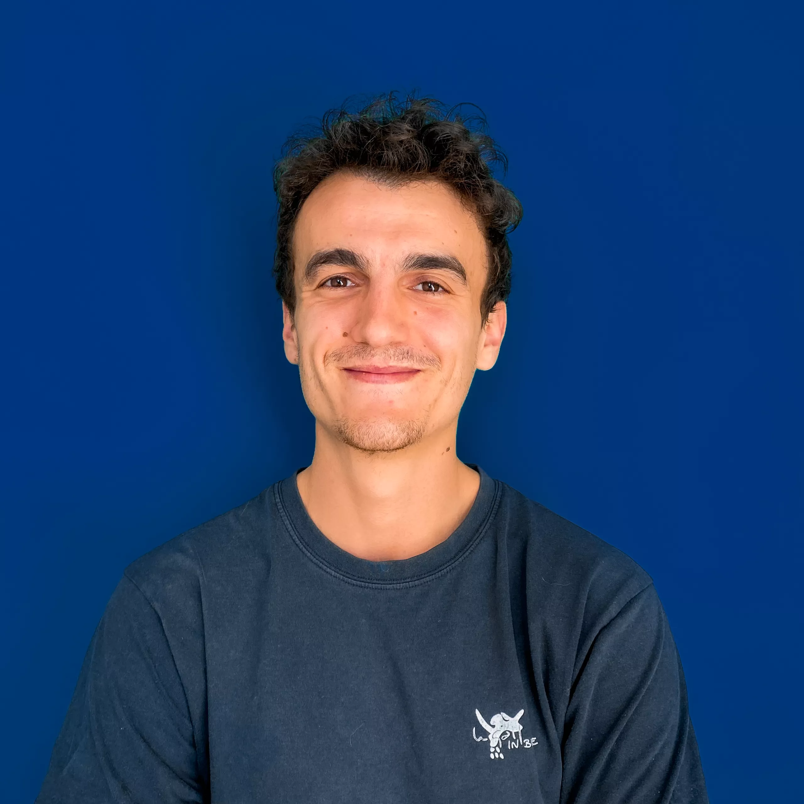 A young man with a slight smile, wearing a dark t-shirt, standing against a solid blue background.