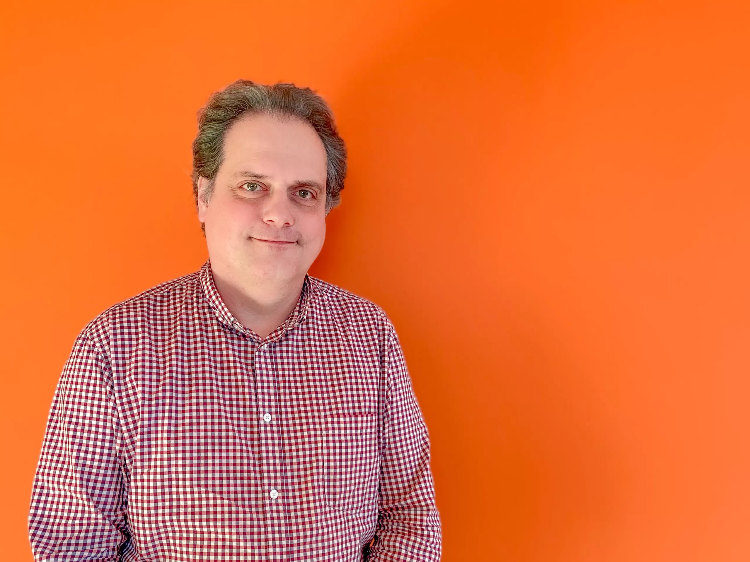 Man in a plaid shirt smiling, standing against a bright orange background.