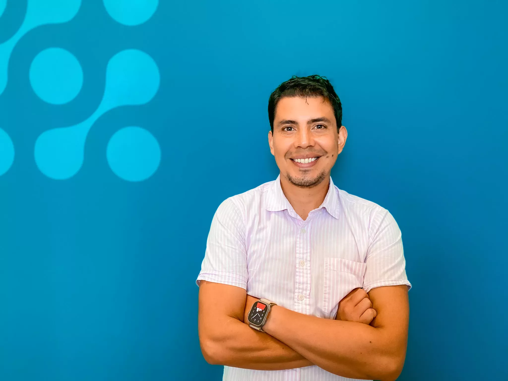 Man in a light striped shirt smiling with arms crossed against a teal background with blue circle patterns.
