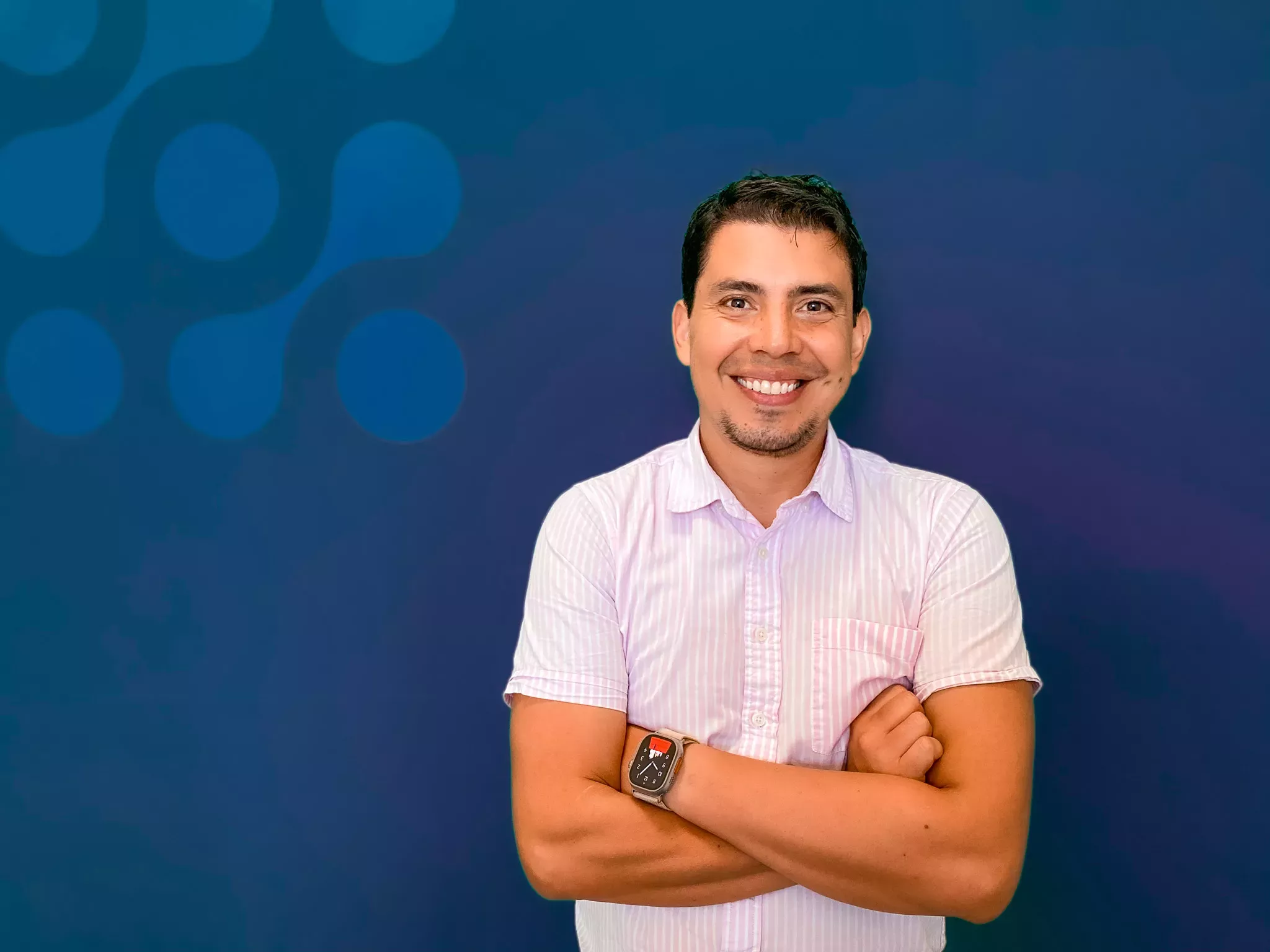 A man smiling at the camera with crossed arms, wearing a striped shirt and a watch, against a blue background with light circles.
