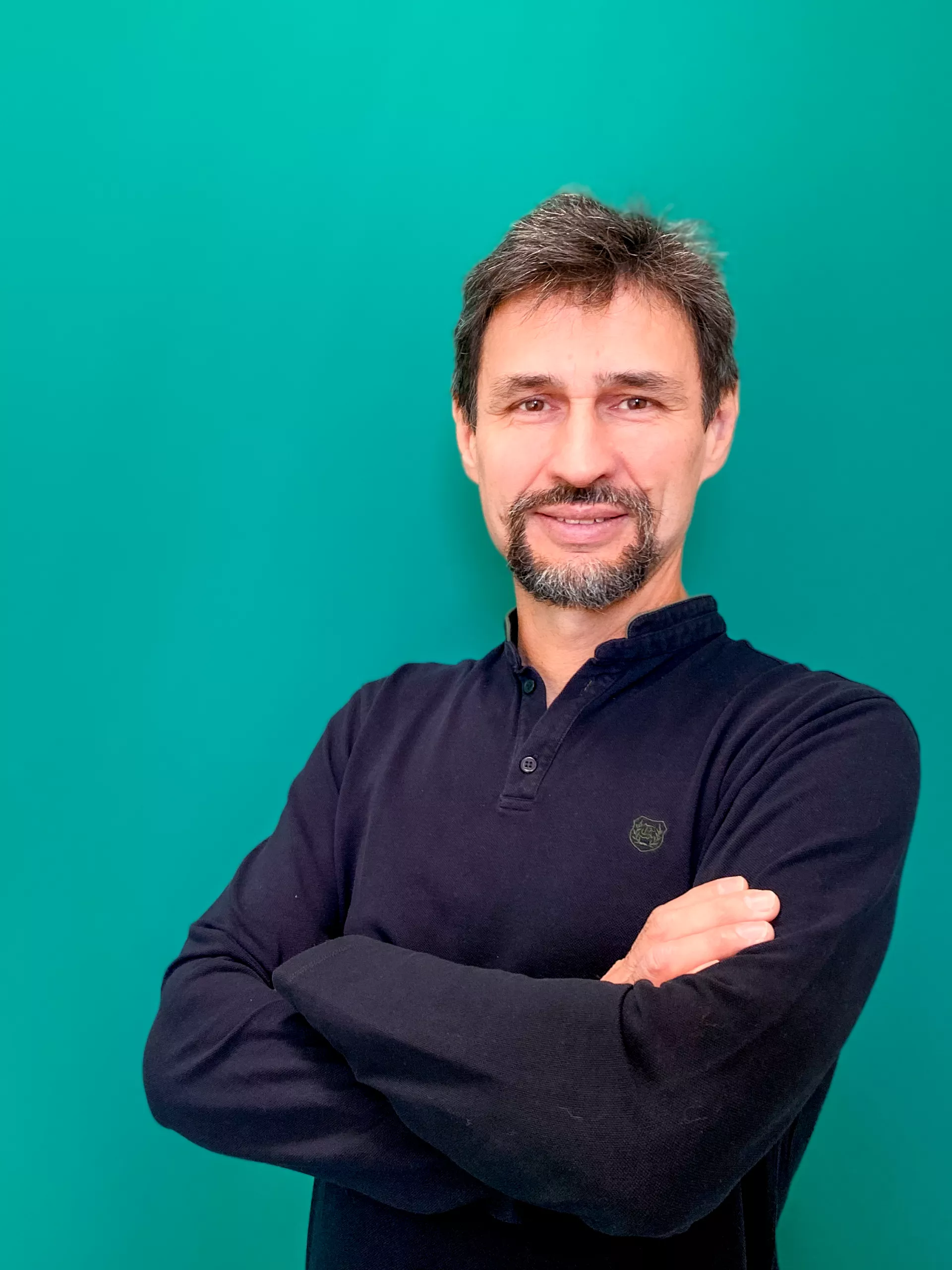 A man with salt and pepper hair, smiling at the camera, arms crossed, wearing a dark polo shirt and standing against a bright green background.