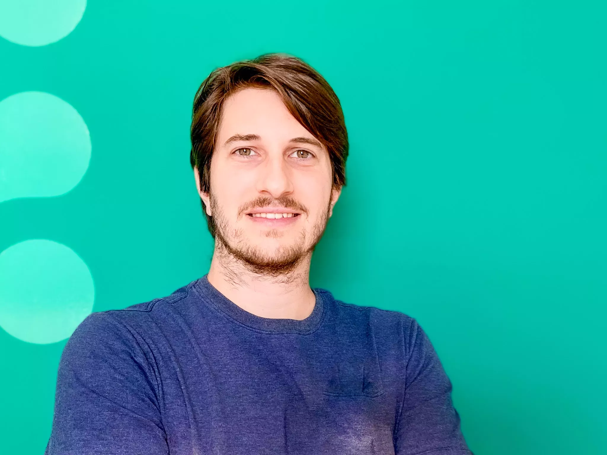 A smiling man with dark hair wearing a blue shirt, against a bright green background with soft white circles.