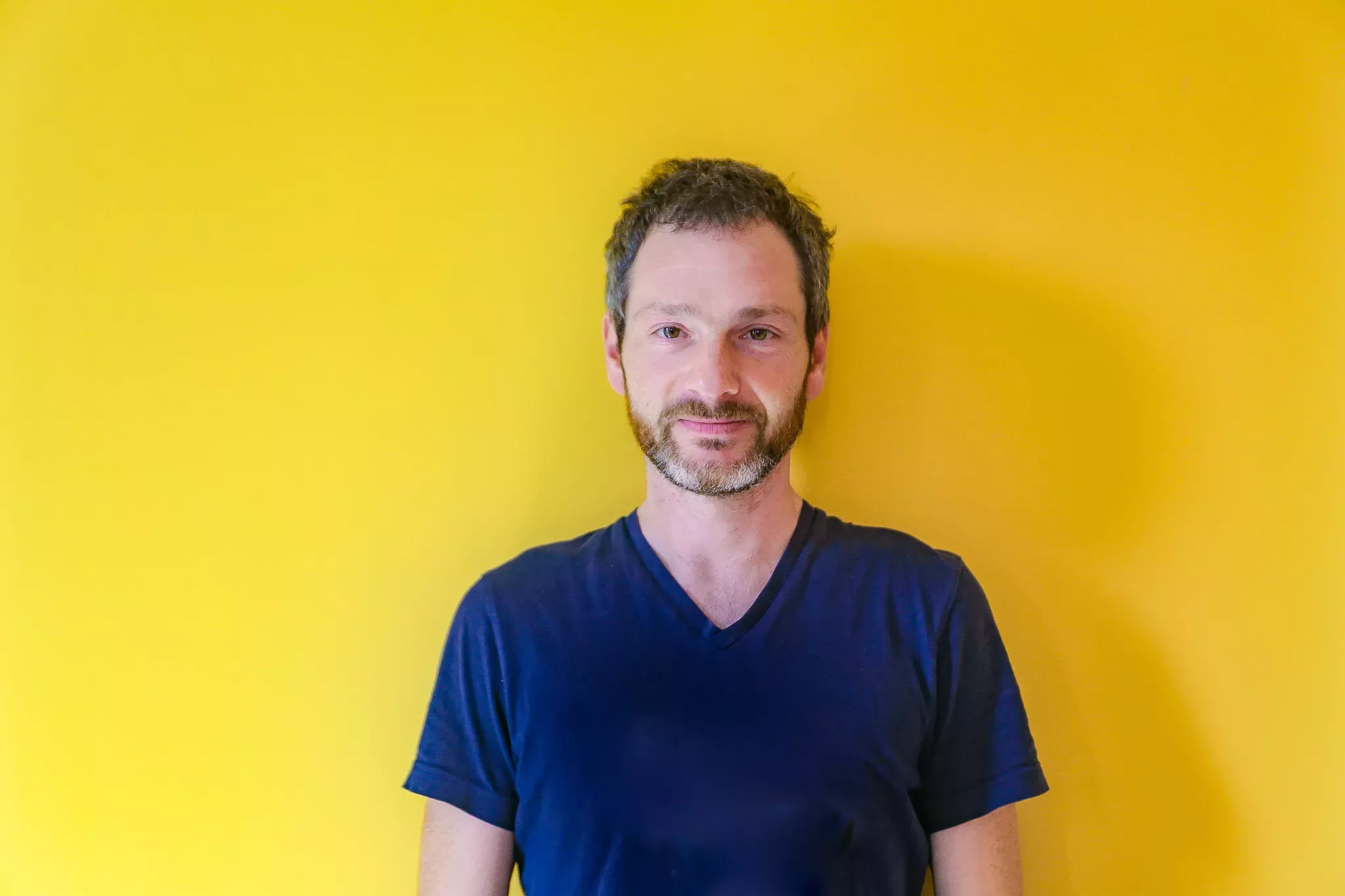 A smiling man with stubble, wearing a blue shirt, stands against a bright yellow background.