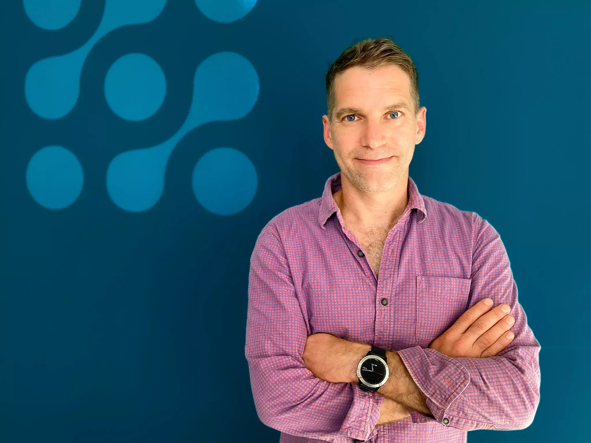 Man in a pink shirt standing with crossed arms, smiling against a blue background with decorative circles.