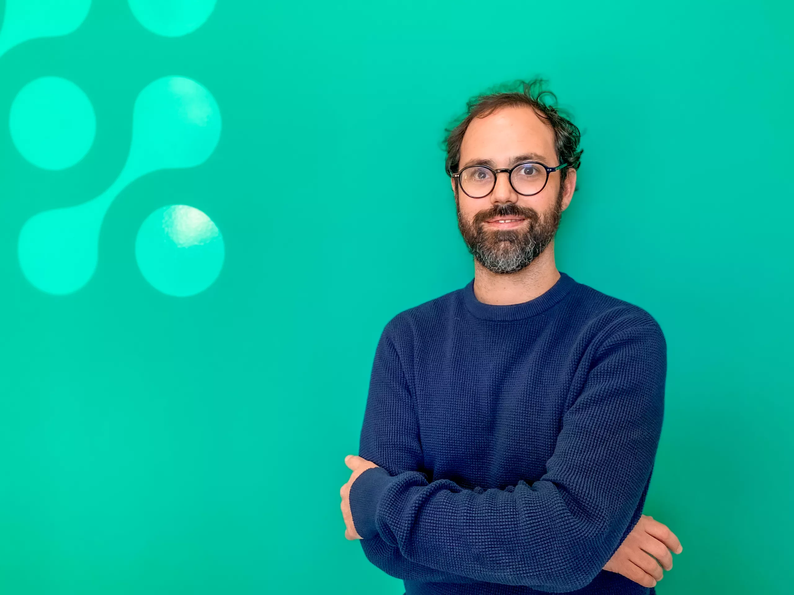 A man with glasses and a beard, wearing a blue sweater, stands with crossed arms against a green background with white bubble shapes.