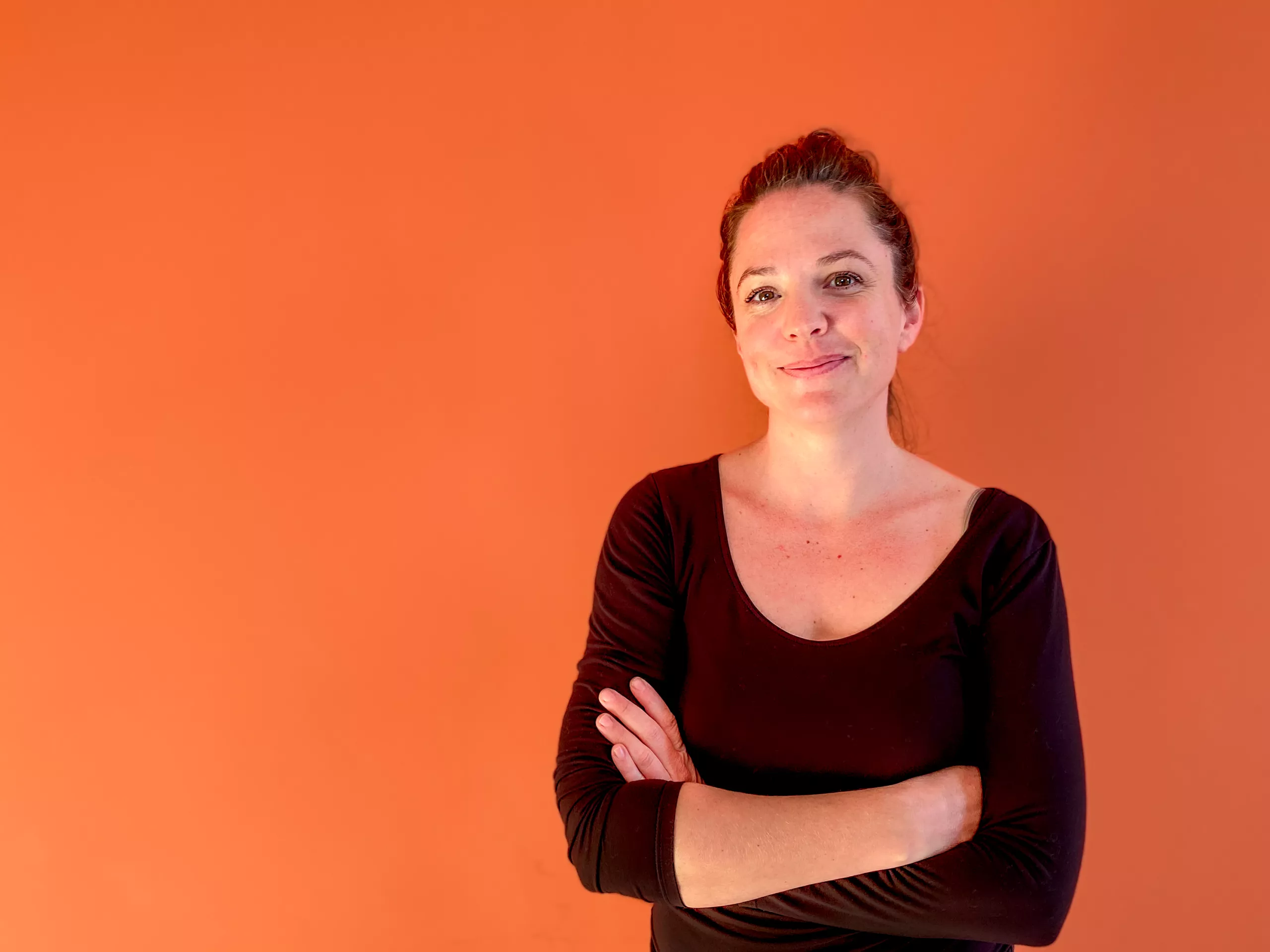 Woman smiling with arms crossed, standing against a solid orange background. she wears a black top and her hair is pulled back.