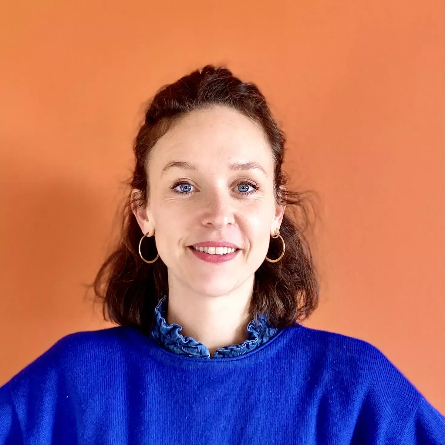 Young woman with blue eyes and brown hair smiling against an orange background, wearing a blue sweater and hoop earrings.