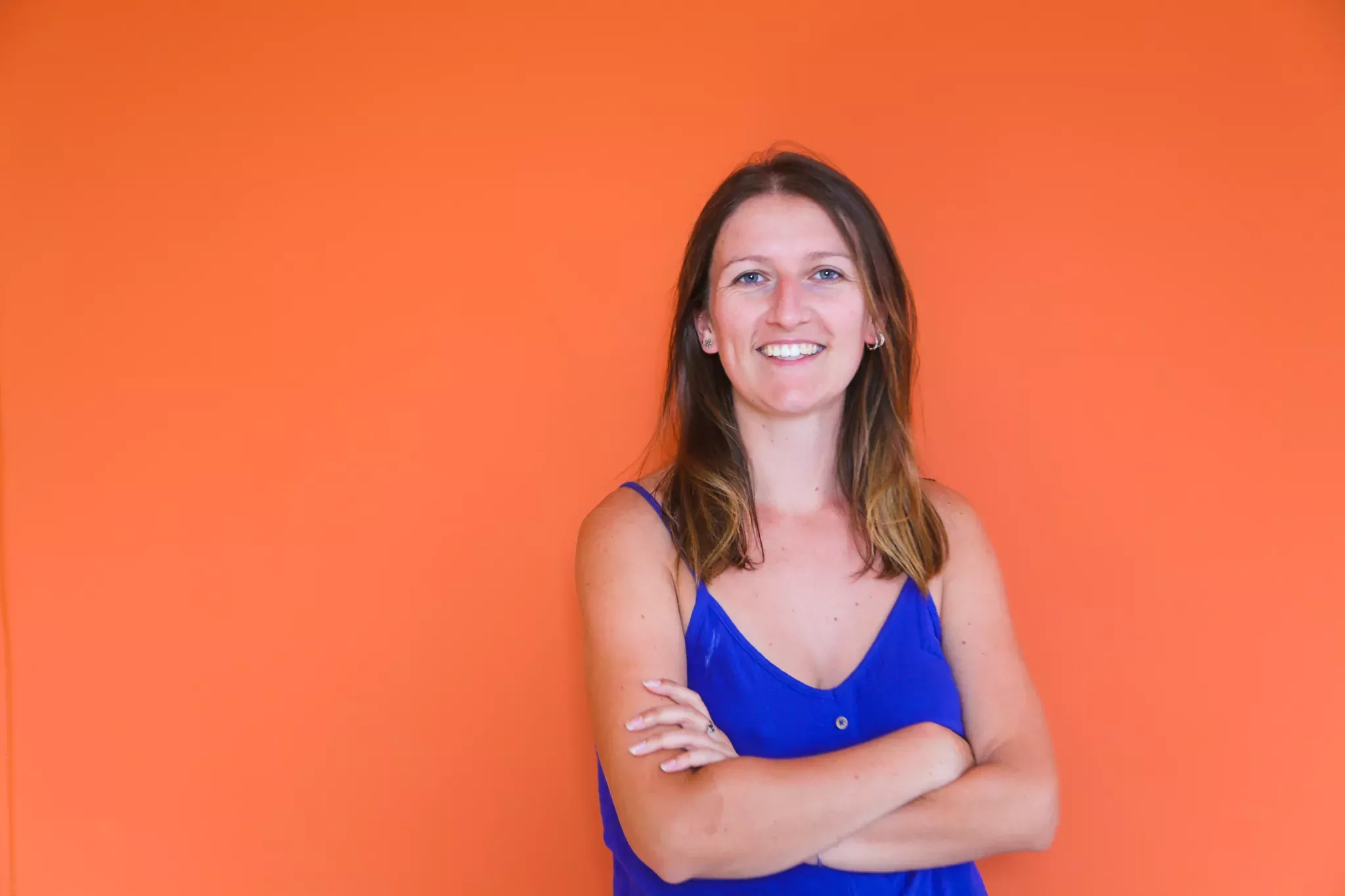 A smiling woman with shoulder-length hair, dressed in a blue top, standing with crossed arms against an orange background.