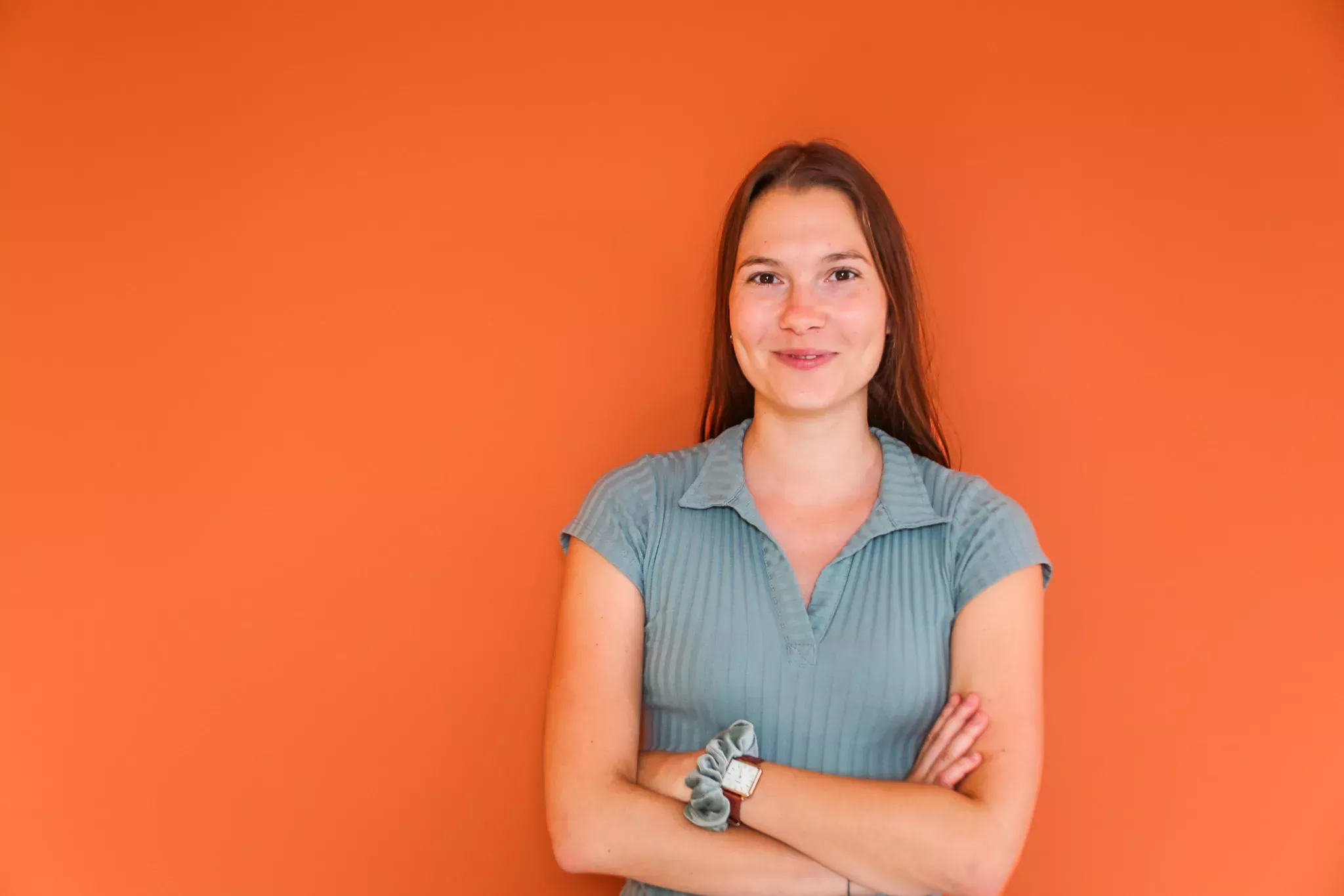 A young woman smiling at the camera, arms crossed, wearing a teal shirt against a bright orange background.