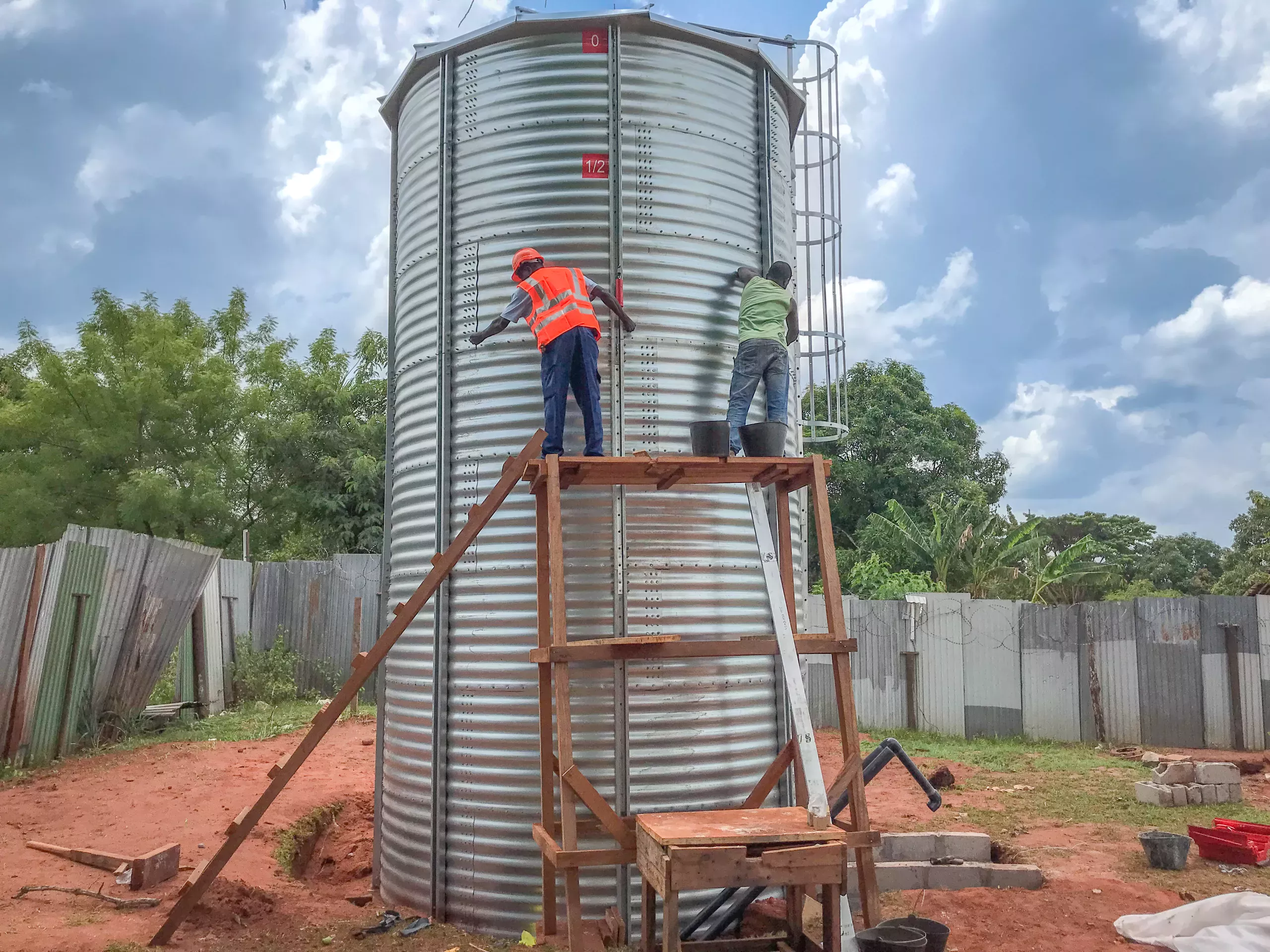 Two workers on a makeshift scaffold performing maintenance on a large metal water tank.