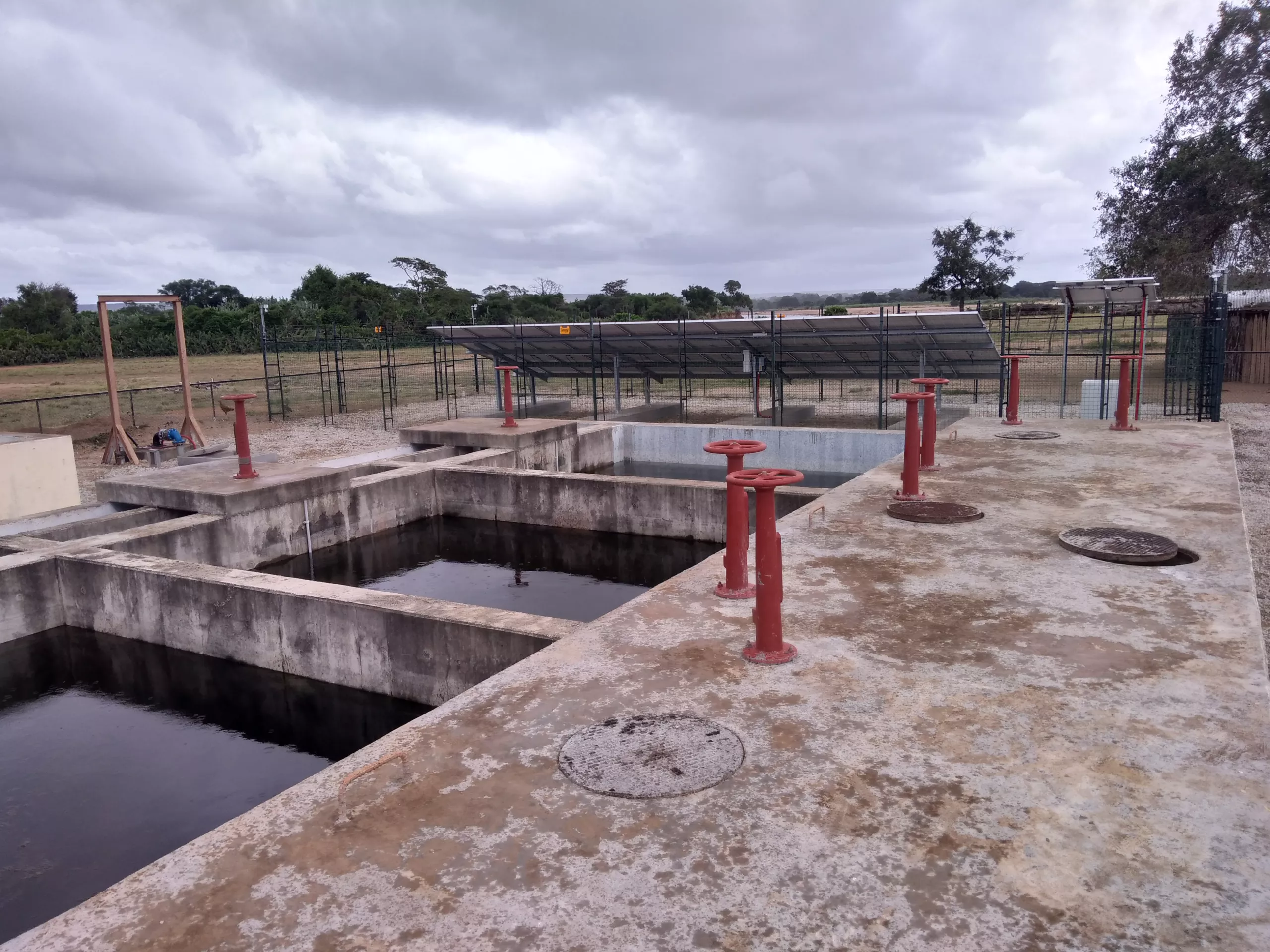 An outdoor water treatment facility in Madagascar features concrete tanks and red metal pipes, ensuring eau potable for the community. A solar panel array stands resilient under a cloudy sky, harnessing clean energy.