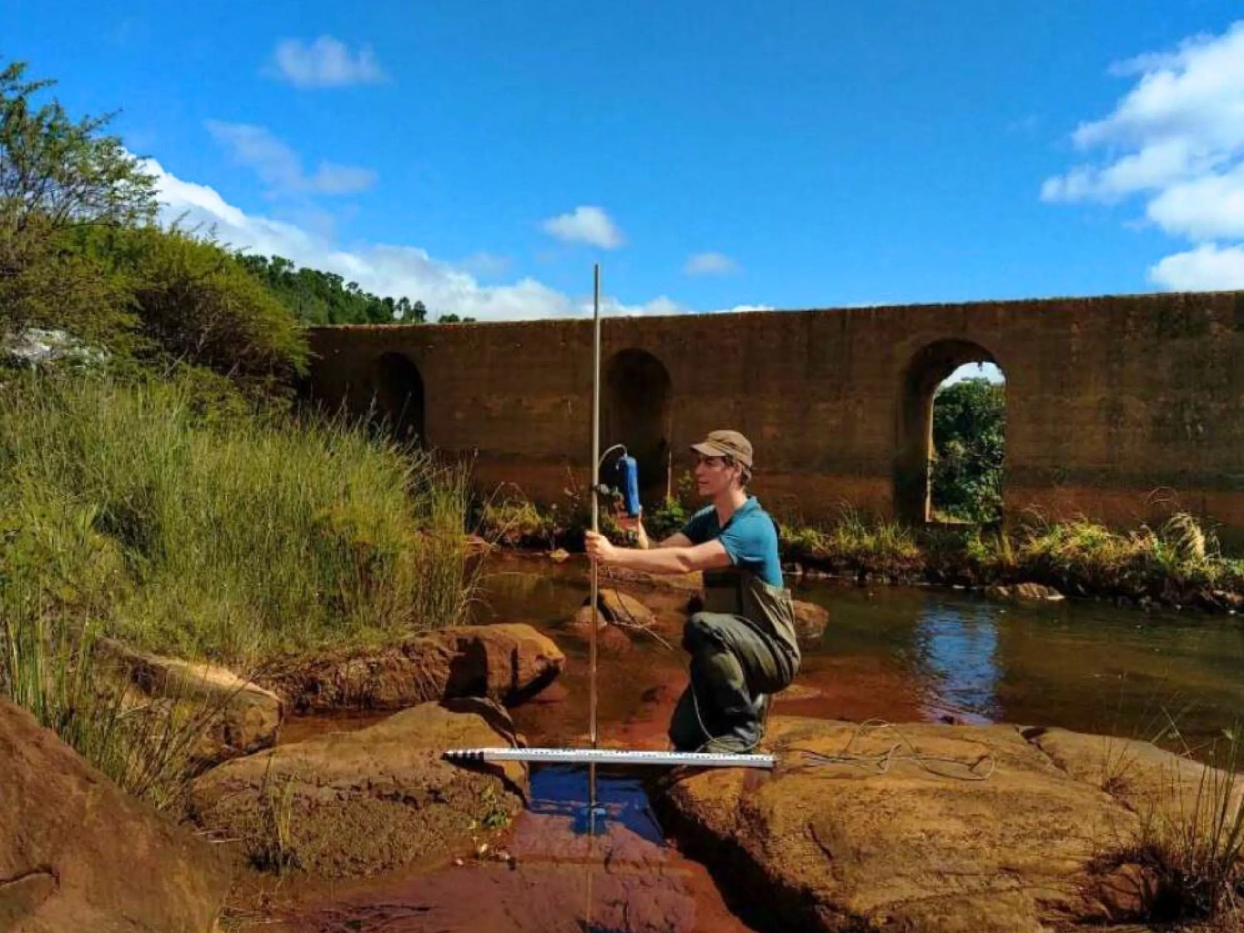 A person kneels on rocks in a shallow stream, holding a large metal frame, with tall grass and an old concrete bridge in the background under a blue sky.