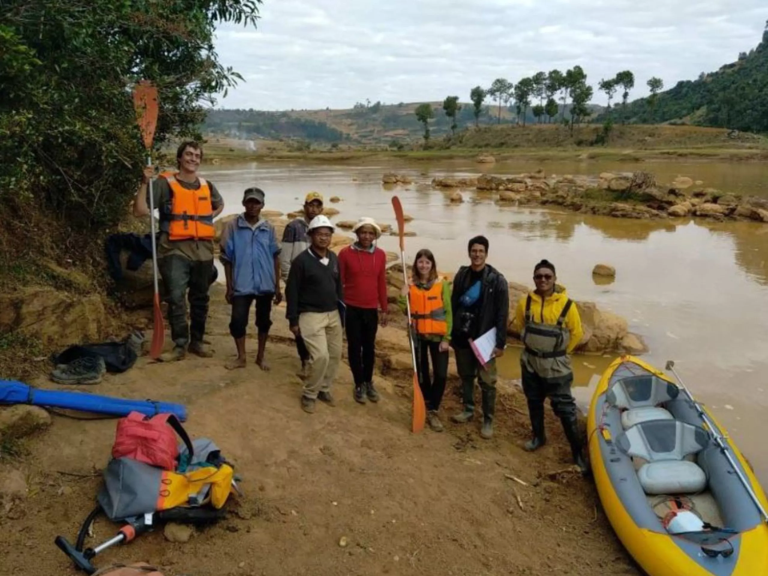 A group of people wearing life jackets and outdoor gear stand by a riverbank next to a yellow inflatable kayak and paddles, with rocky terrain and trees in the background.