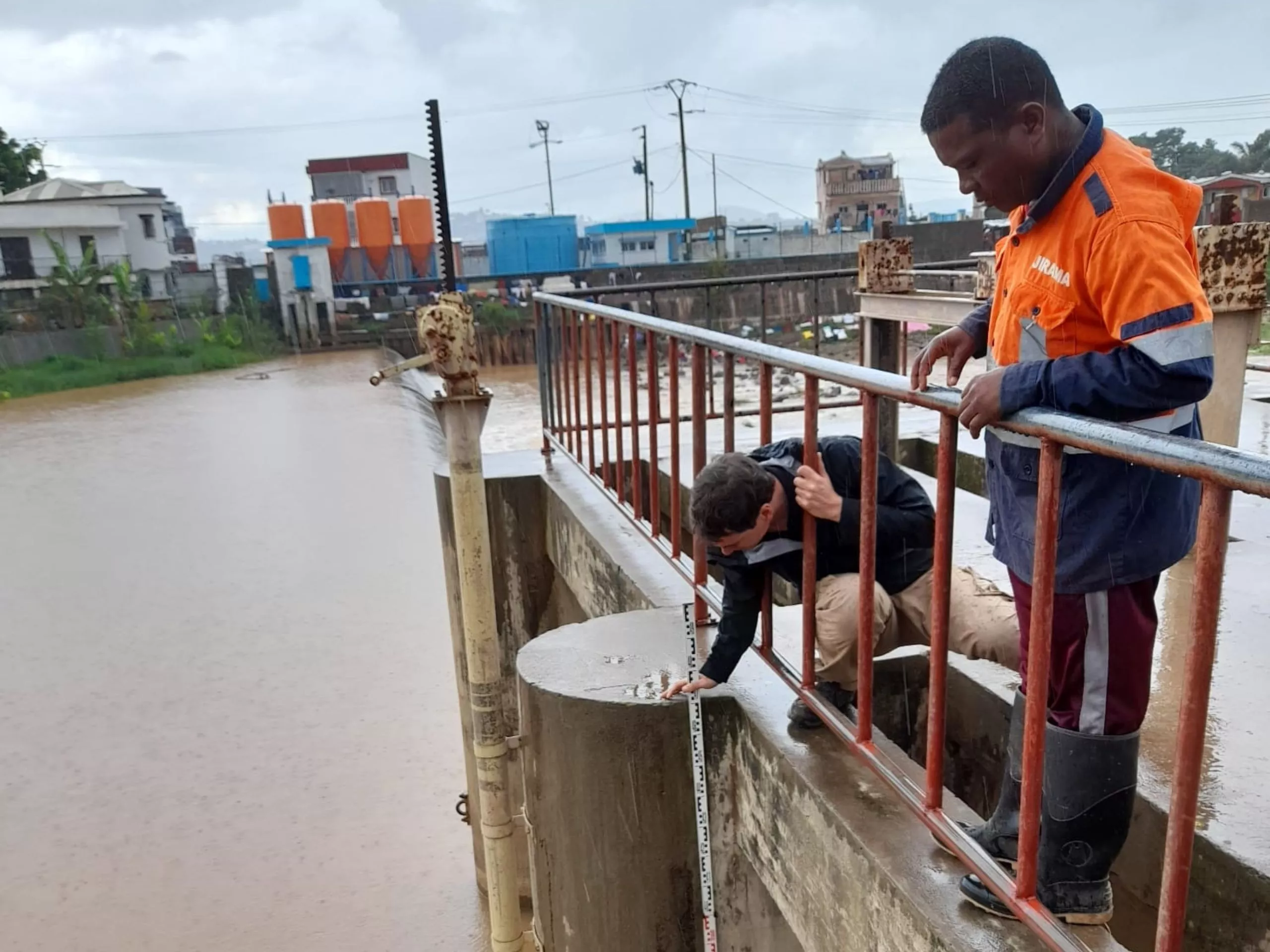 Two people check water levels at the edge of a flooded area during heavy rain, with one person reading a measuring gauge attached to a concrete post.