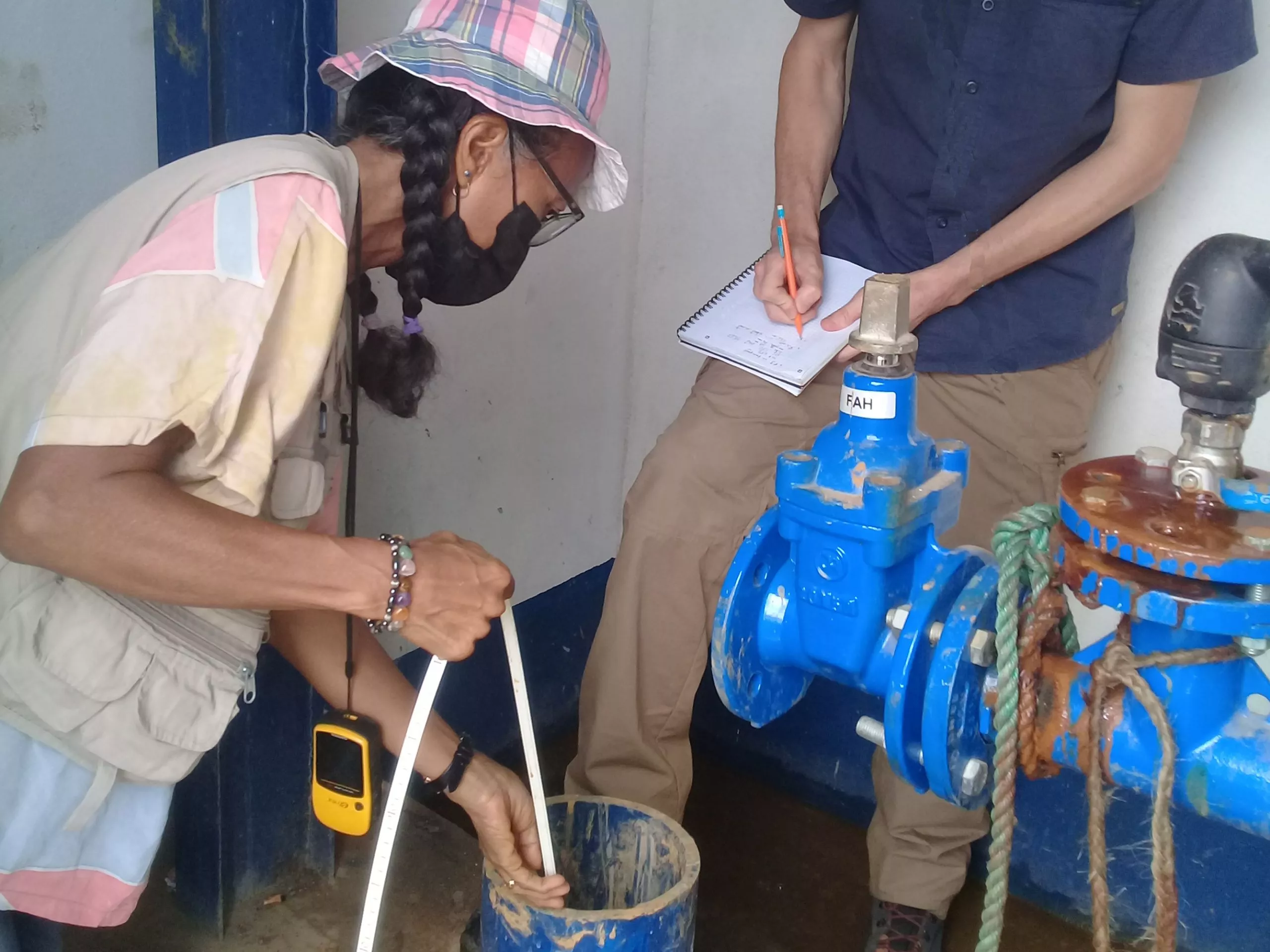 A person collects a water sample in a bucket while another person takes notes beside blue industrial valves.