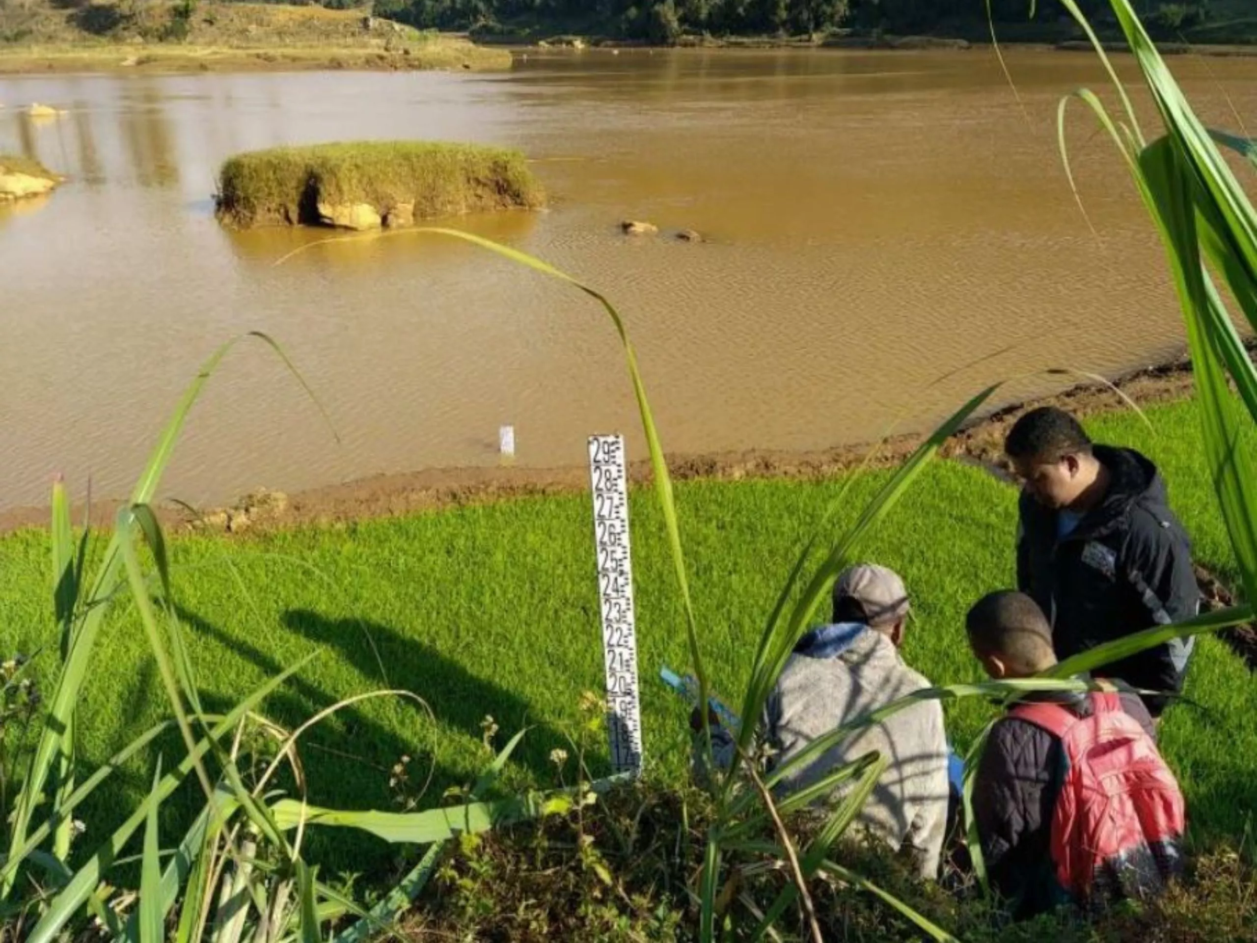 Three people are gathered near a water level gauge by the edge of a river or lake with muddy water and grassy banks.
