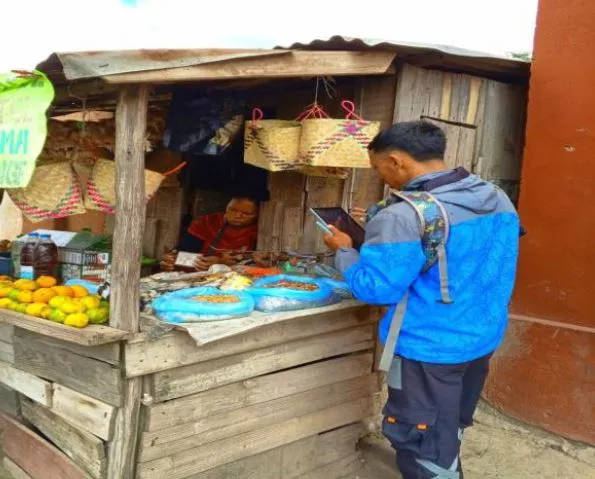 A person in a blue jacket buys food from a wooden roadside stall displaying fruits and snacks, with a vendor inside the stall.