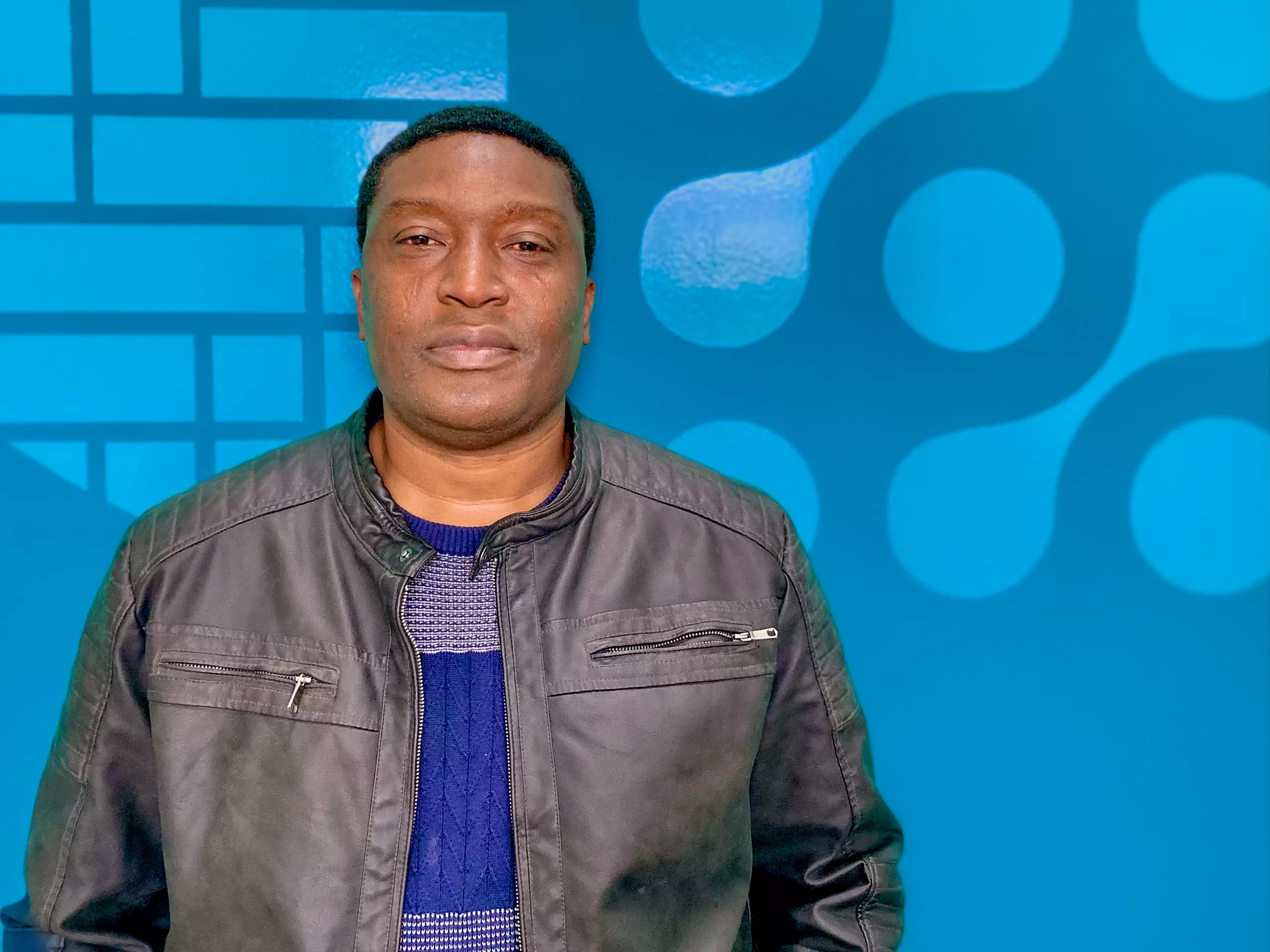 A man wearing a black leather jacket and blue sweater stands in front of a blue background with abstract circular and grid patterns, representing the innovative spirit of the Hydroconseil ingénierie équipe.