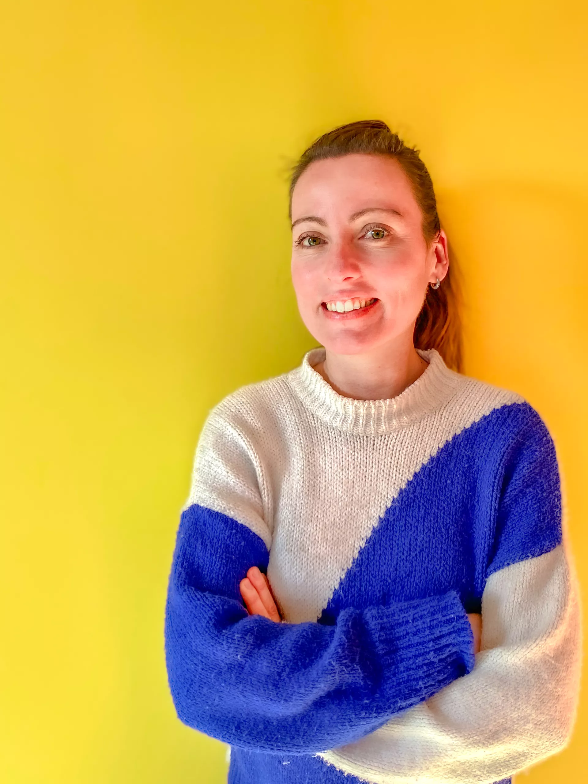 A woman with light skin and brown hair in a ponytail stands against a yellow wall, smiling with arms crossed, wearing a white and blue sweater.