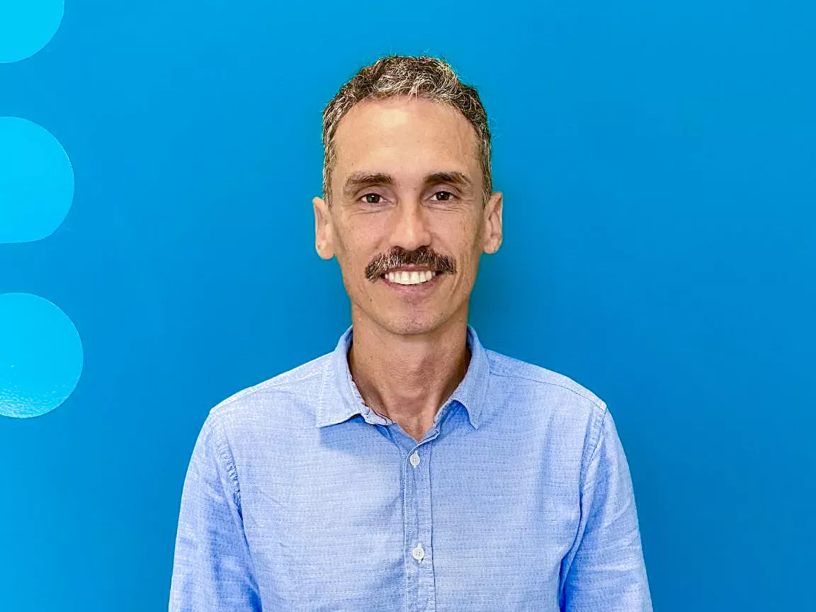 A man with short curly hair and a mustache, wearing a light blue button-up shirt, stands in front of a solid bright blue background.