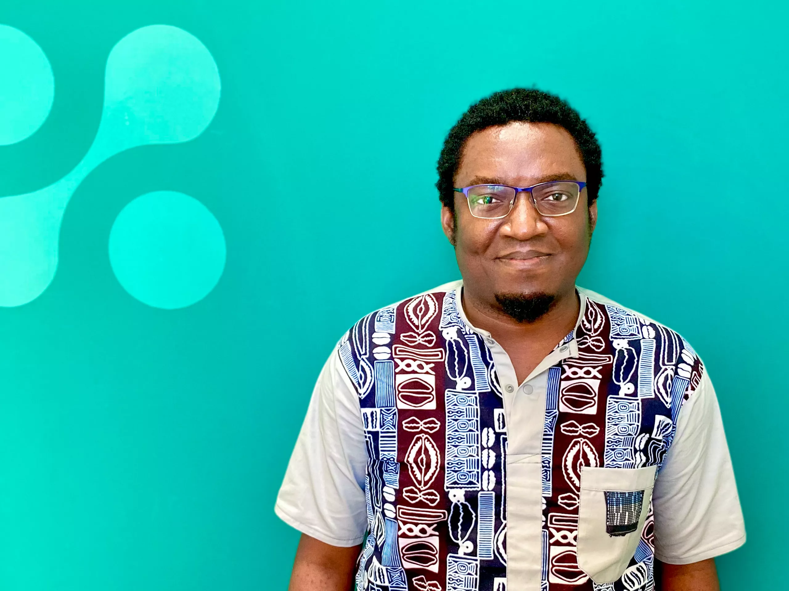 A man wearing glasses and a patterned shirt stands in front of a teal background with circular abstract shapes.