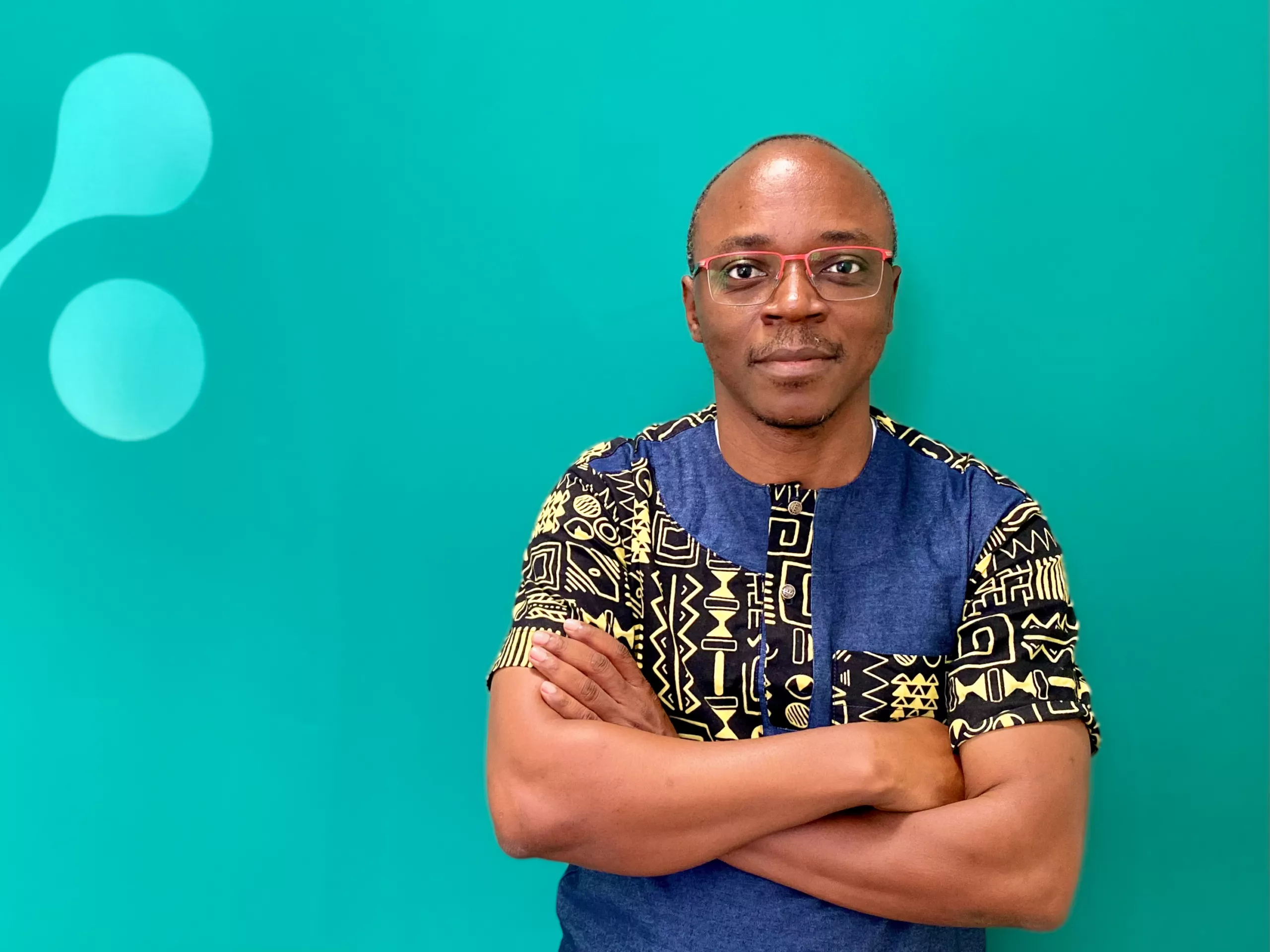 A person wearing glasses and a patterned shirt stands with arms crossed in front of a teal background featuring abstract shapes.