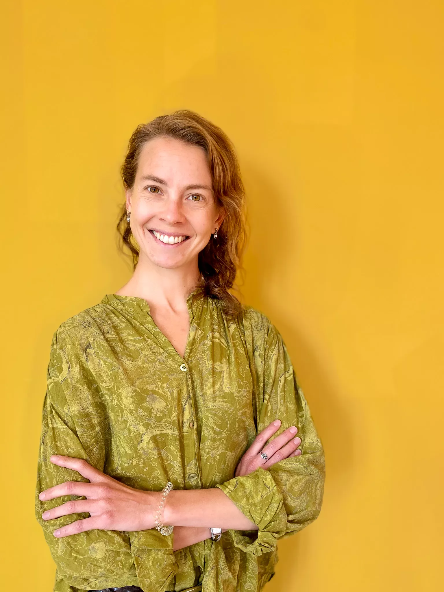 A person stands against a yellow background, smiling, with arms crossed and wearing a green patterned blouse.