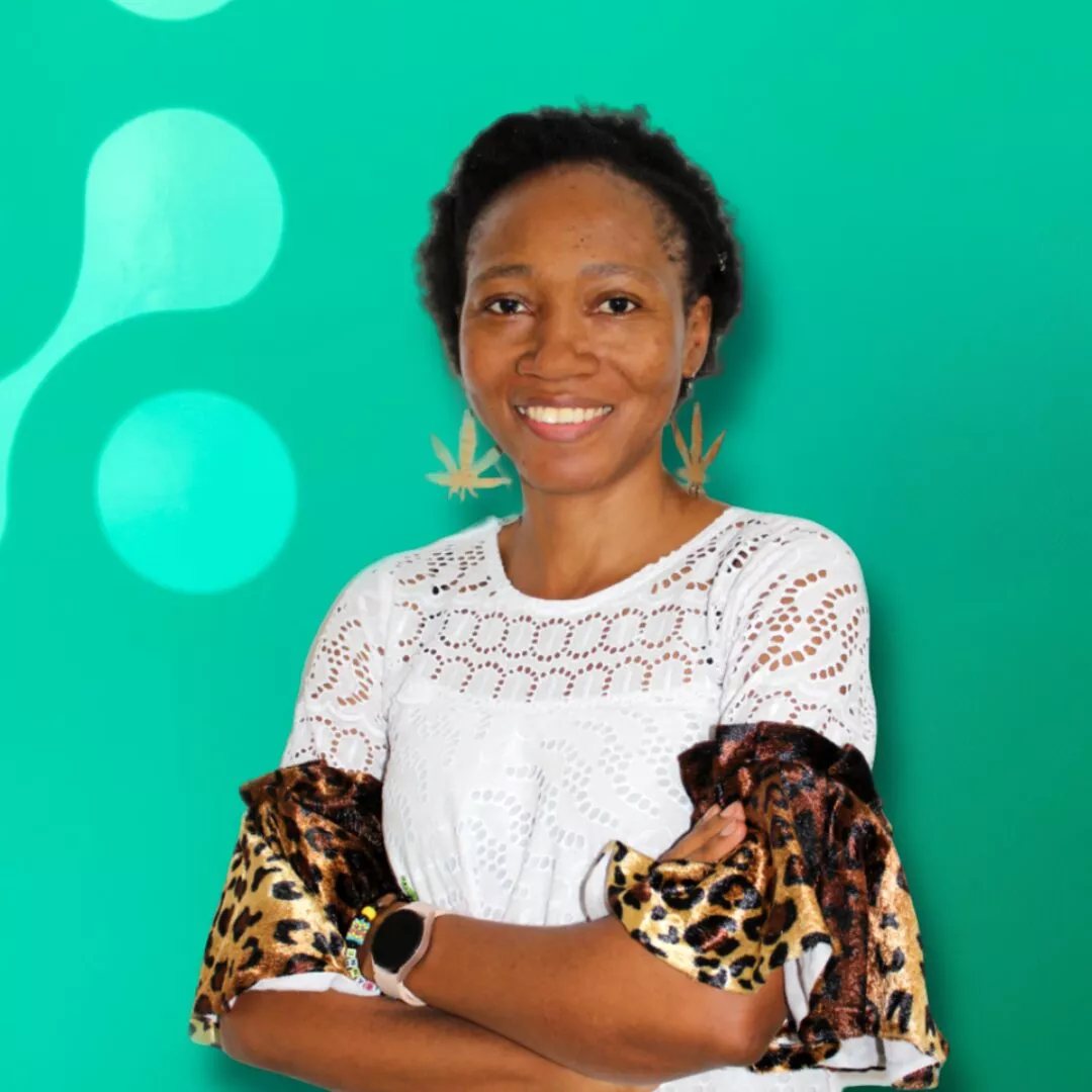A woman with short hair smiles at the camera, wearing a white lace top with leopard print accents and leaf-shaped earrings, standing against a teal background with abstract patterns.