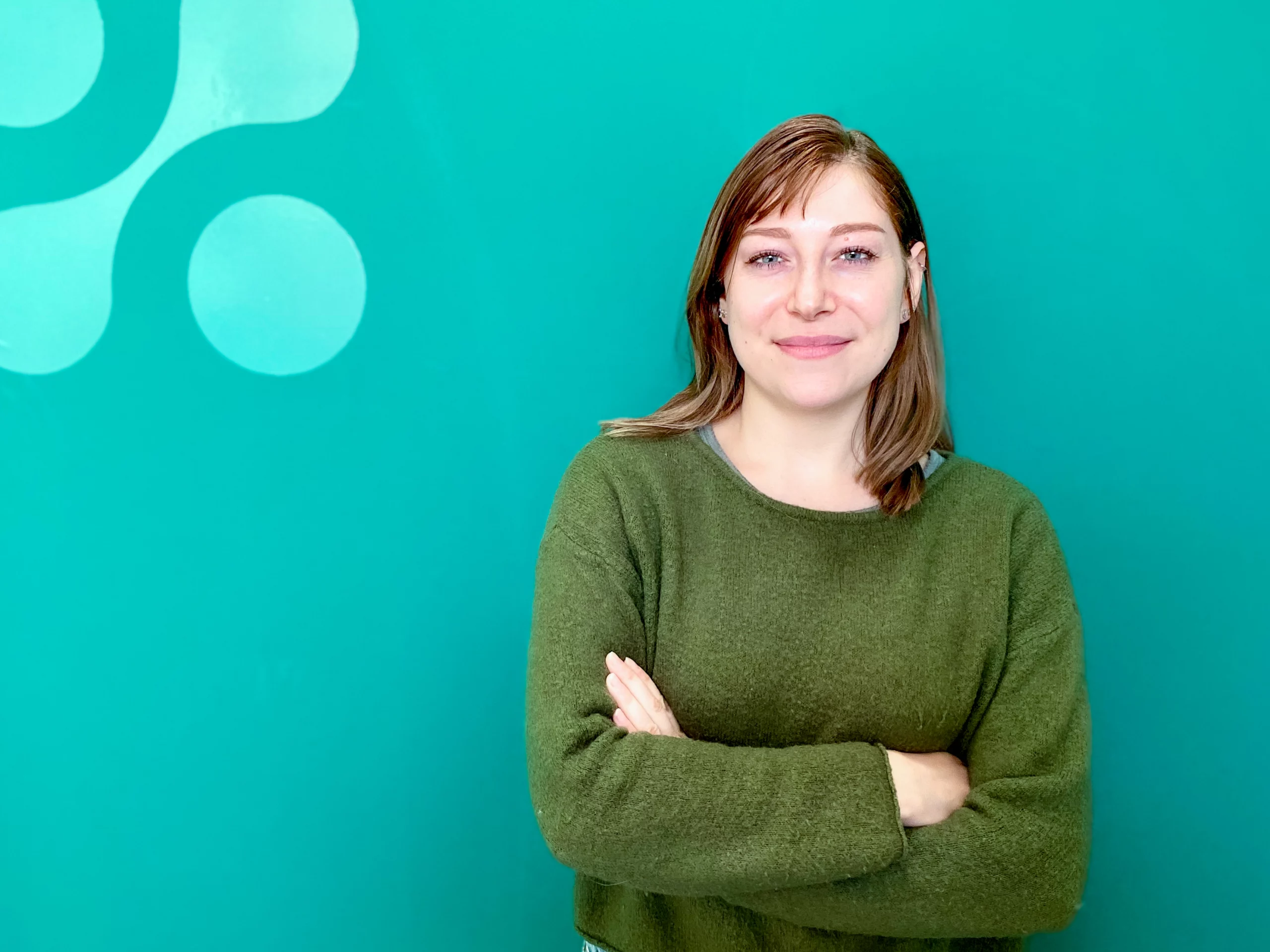 Une femme aux cheveux châtain clair, vêtue d'un pull vert, se tient debout, les bras croisés, devant un mur bleu sarcelle orné de motifs circulaires.