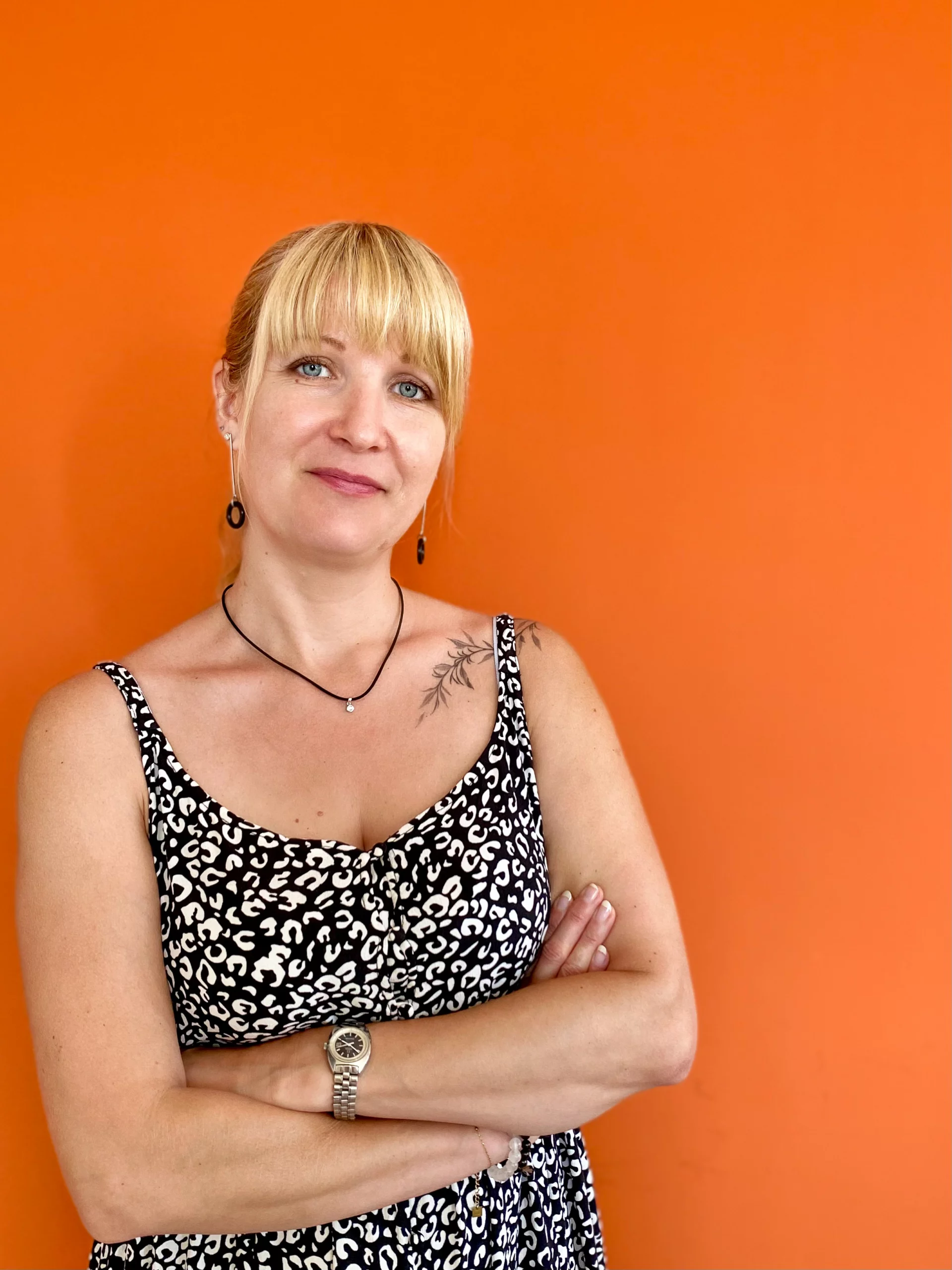 Une femme aux cheveux blonds, arborant un tatouage sur l'épaule gauche, se tient debout, les bras croisés, devant un fond orange, vêtue d'une robe sans manches à motifs.