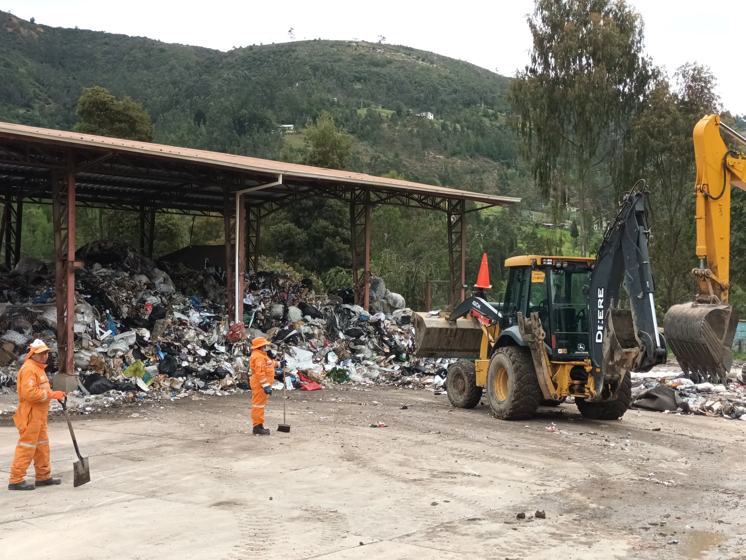 Deux ouvriers vêtus de combinaisons orange nettoient à proximité d'une chargeuse-pelleteuse sur une décharge couverte où s'entassent des déchets ; on aperçoit des collines verdoyantes en arrière-plan.