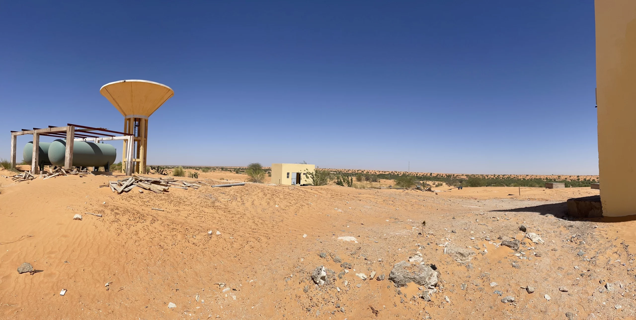 Une scène de désert sablonneux avec un grand réservoir d'eau sur pilotis, un réservoir cylindrique plus petit probablement utilisé pour l'assainissement, des débris éparpillés et un petit bâtiment jaune à l'arrière-plan, sous un ciel bleu limpide.