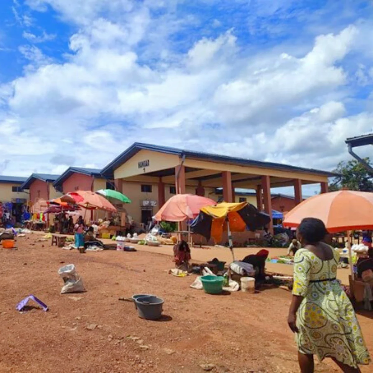 Scène de marché en plein air avec plusieurs personnes, des parasols colorés, des marchandises éparpillées et un bâtiment portant l'inscription « Market » sous un ciel partiellement nuageux.