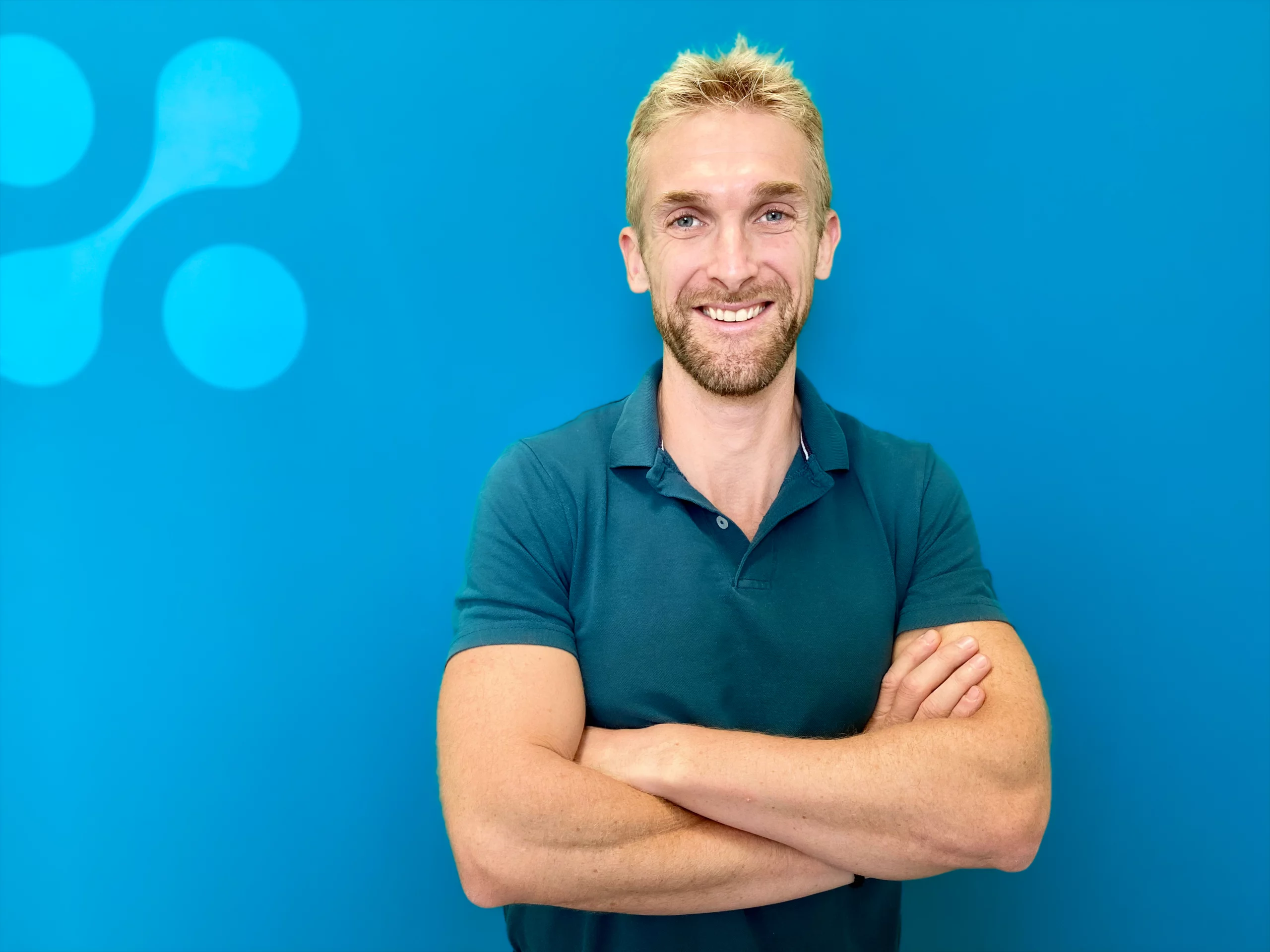 Un homme souriant, barbu, aux cheveux blonds courts, vêtu d'un polo bleu sarcelle, se tient debout, les bras croisés, devant un mur bleu.